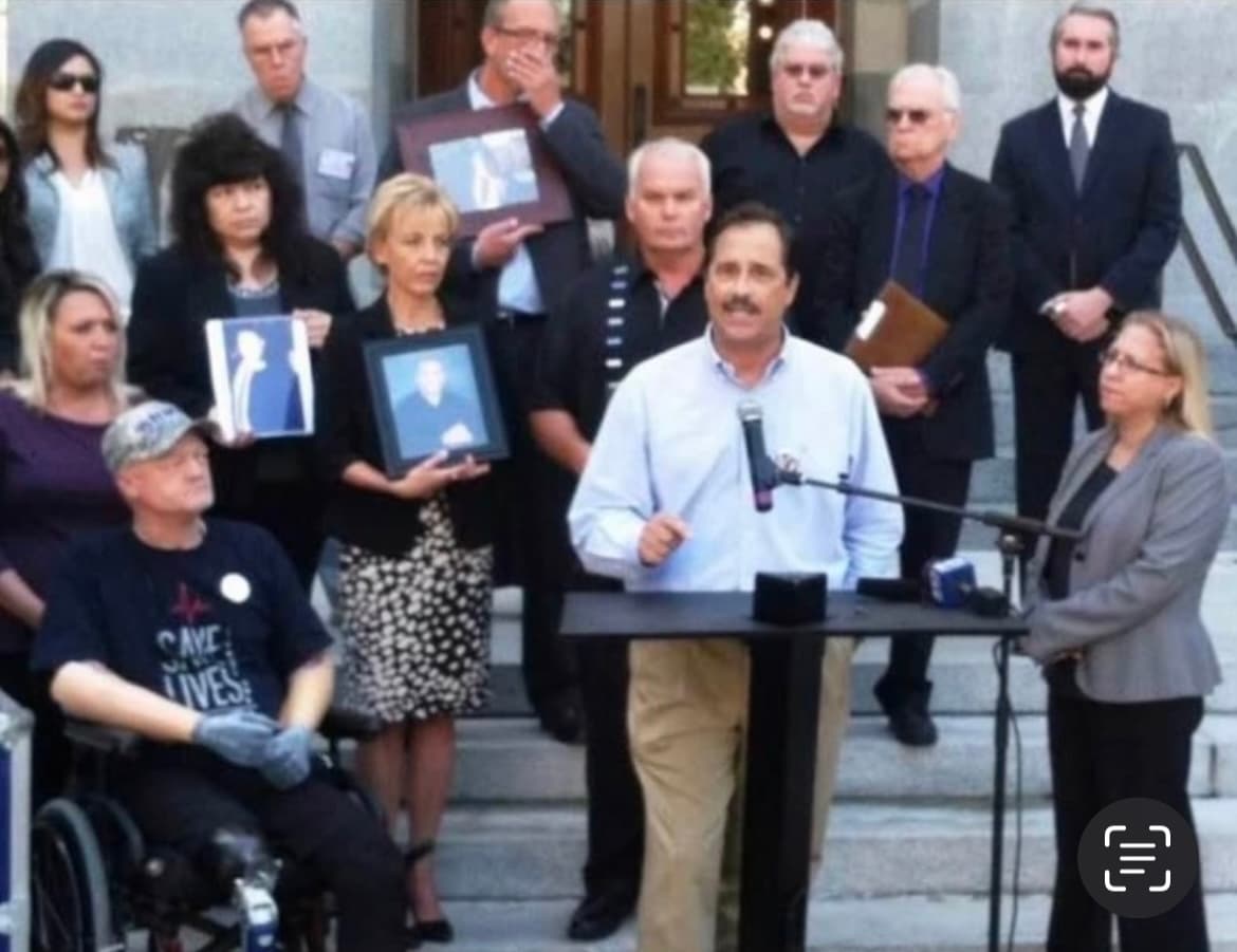 Bob Pack speaking at a podium at an outdoor advocacy event, with supporters and community members